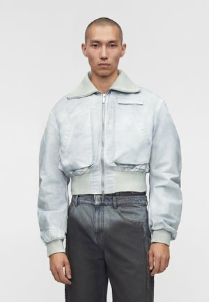 Άνδρας που φοράει ανοιχτό γκρι cropped bomber μπουφάν με φερμουάρ μπροστά και ψηλόμεσα σκούρα γκρι τζιν μπροστά σε απλό φόντο.
