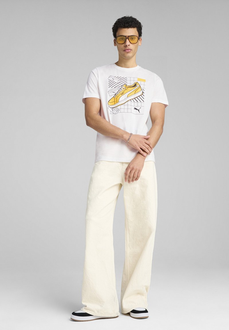 Camiseta blanca con gráfico de zapatillas amarillas y contornos negros. Combinada con pantalones sueltos de color crema y zapatos en blanco y negro.