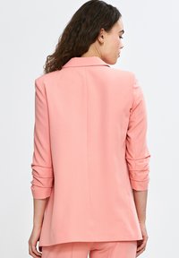Różowa marynarka o dopasowanym kroju, z długimi rękawami podwiniętymi na mankietach i jednym rozcięciem z tyłu. Gładki materiał o lekko teksturowanej powierzchni.