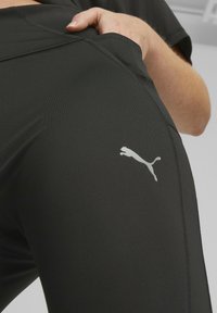 Czarne legginsy sportowe wykonane z elastycznego materiału, charakteryzujące się dopasowanym krojem i odblaskowym szarym logo Pumy na udzie.