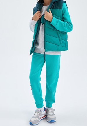 Barn iført turkis sweatpants med grafik, matchende puffervest, grå sweatshirt med hjerte, hvide sokker og grå sneakers med velcrolukning.