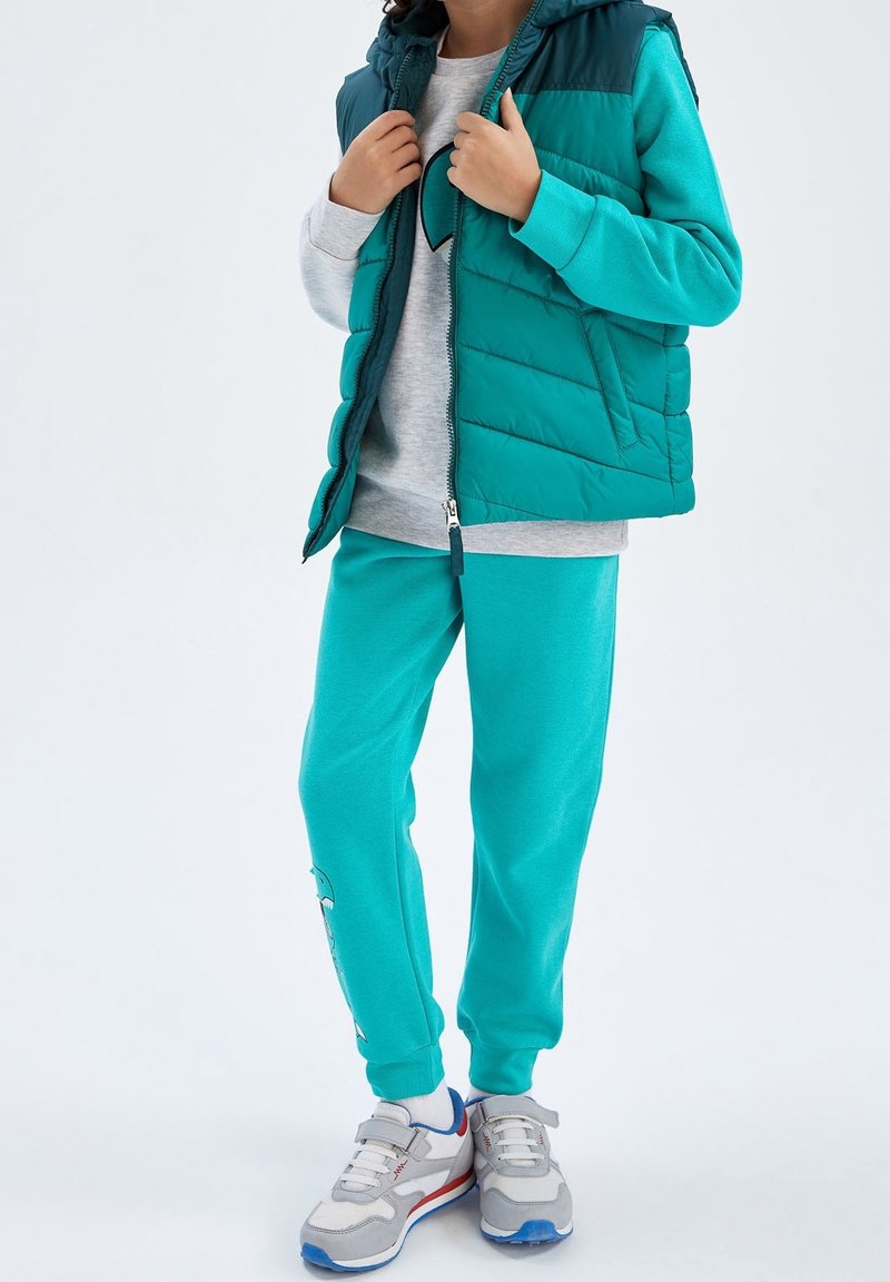 Enfant portant un pantalon de survêtement turquoise avec un motif, une doudoune assortie, un sweat-shirt gris avec un cœur, des chaussettes blanches et des baskets grises à bandes velcro.