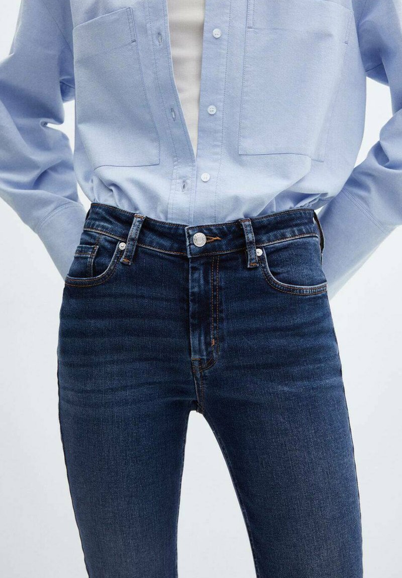 Jean taille haute en denim foncé avec une texture lisse, un délavage subtil, un design classique à cinq poches et des coutures contrastées. Associé à une chemise à boutons bleu clair.