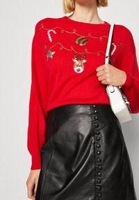 Rode gebreide trui met feestelijke versieringen; gecombineerd met een zwarte leren rok met een knoop zijdetail. Model draagt een witte tas.