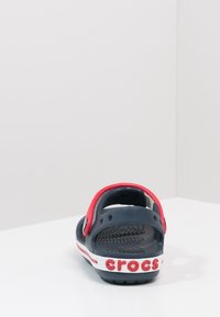 Granatowe Crocsy z czerwoną paskiem na pięcie, z wentylacyjnymi otworami i białą podeszwą z czerwoną marką "crocs". Teksturowane wnętrze.