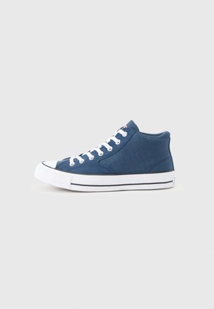 Blauwe hoge canvas sneaker met witte veters en rubberen zool, zijaanzicht op een eenvoudige lichtgrijze achtergrond.