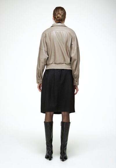 Femme vue de dos portant une veste en cuir taupe, une jupe noire mi-genoux, des collants transparents et des bottes à talons noires montant jusqu'aux genoux.