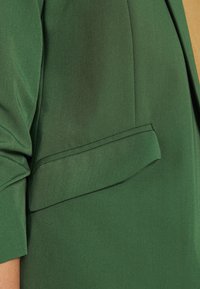 Gros plan sur la manche d'une veste verte et une poche avec rabat, montrant la texture du tissu et les détails de la couture.