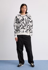 Fekete-fehér kötött pulóver, absztrakt mintával, bordás mandzsettákkal és aljjal, laza fekete nadrággal és fekete sportcipővel párosítva.