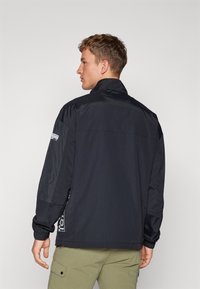 Chaqueta cortavientos negra con cuello alto, puños elásticos y detalle de logo en el lado, diseñada con una textura suave y brillante.