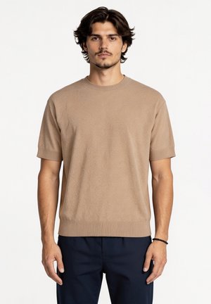 Mężczyzna o ciemnych włosach noszący beżowy krótki sweter z dzianiny i ciemnoniebieskie spodnie, stojący na jednolitym białym tle.