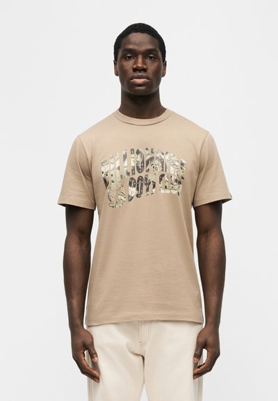 Άνδρας που φοράει μπεζ μπλουζάκι με το λογότυπο "Billionaire Boys Club" και ανοιχτόχρωμο παντελόνι, στέκεται μπροστά σε ένα απλό λευκό φόντο.