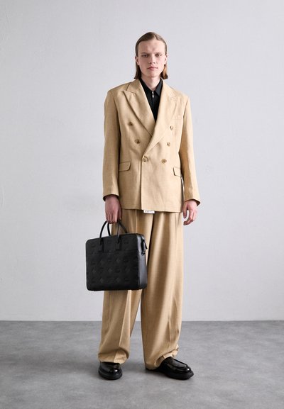 Blazer beige à double boutonnage, pantalon beige à jambes larges, chemise noire et chaussures noires. Sac noir à motif avec double anse, sur un fond neutre.