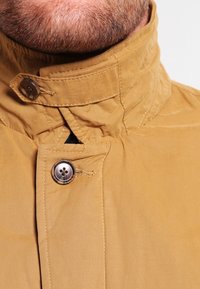 Beige, lätt jacka med hög krage, som har två knappar, stickningar som accent och en slät, något strukturerad yta.