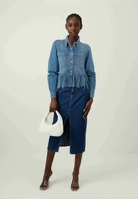 Camisa de denim com cintura peplum, dois bolsos no peito; combinada com uma saia longa e reta de denim com uma fenda. A segurar uma mala acolchoada branca.