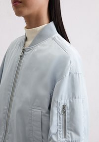 Jasnoniebieska kurtka bomberka z prążkowanym kołnierzem, zamykanymi na zamek kieszeniami i gładką teksturą. Posiada kieszenie po bokach oraz kieszeń na ramieniu zapinaną na zamek.