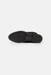 Suela de zapato negra con un patrón de agarre texturizado y circular, y un pequeño área de logo rectangular sobre un fondo blanco liso.