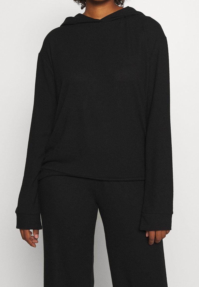 Personne portant un sweat à capuche noir à manches longues et un pantalon noir assorti sur un fond clair uni.