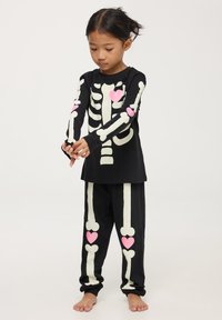Set di pigiama a maniche lunghe nero con stampa scheletro che si illumina al buio e dettagli a forma di cuore rosa. Tessuto morbido con vestibilità comoda.