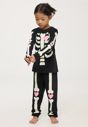 Set di pigiama a maniche lunghe nero con stampa scheletro che si illumina al buio e dettagli a forma di cuore rosa. Tessuto morbido con vestibilità comoda.