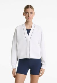 Modelka stoi pewnie, ubrana w białą, skrojoną kurtkę z modnymi detalami na granatowym body i szortach, na neutralnym tle.