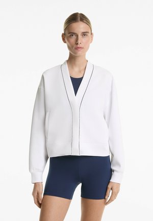 Model stoji samouvjereno, odjevena u bijelu skraćenu jaknu s elegantnim detaljima preko mornarsko plave bodi haljine i šortseva, pred neutralnom pozadinom.