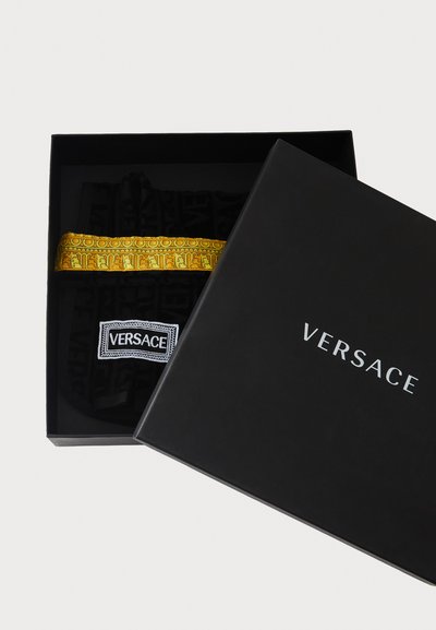 Ținută neagră Versace cu bandă cu model auriu în interior, într-o cutie neagră deschisă, cu sigla "Versace" pe capac.
