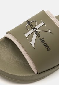 Grønn gummisandal med en beige teksturert stropp, med preget svart logo "Calvin Klein Jeans" på fronten.