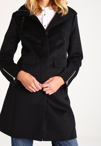 Cappotto nero con un design strutturato, polsini con zip e due tasche frontali. Presenta un ampio colletto e una silhouette aderente.