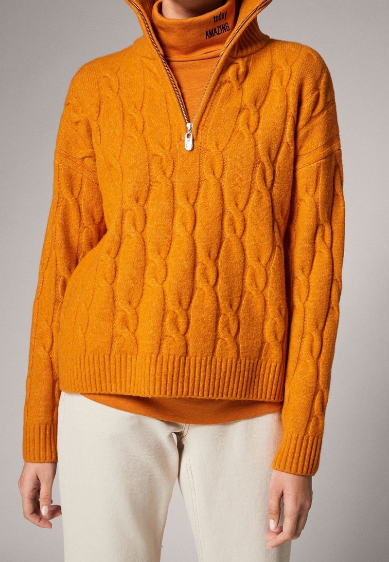 Maglione arancione a maglia a trecce con un collo alto zippato e orlo a coste, indossato sopra un dolcevita abbinato. Texture morbida, vestibilità casual.