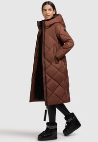 Brūns, garš quilted pūkains mētelis ar kapuci, ar priekšējo rāvējslēdzēju, sānu kabatām un melnu oderi. Melni sniega zābaki papildina apģērbu.