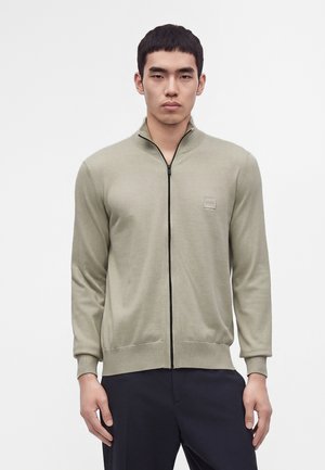 Young man wearing a light beige zip-up sweater with a small logo on the chest and dark pants, standing against a plain background.