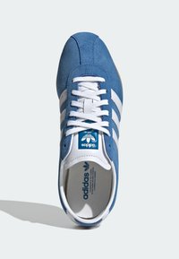 Scarpa sportiva in camoscio blu con strisce bianche, lacci bianchi e un'etichetta blu all'interno. Caratterizzata da una punta arrotondata e una soletta testurizzata.