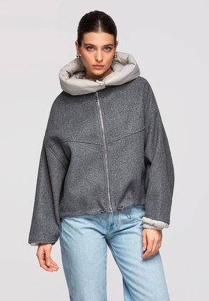 Kvinde iført en grå lynlåstrøje med en stor lysegrå polstret krave og lyseblå jeans, poserer foran en ensfarvet baggrund.