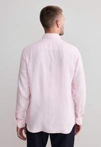 Lichtroze linnen overhemd met lange mouwen, kleine knopen en een afgeronde zoom, gezien van achteren. Kenmerkt zich door een simpel ontwerp en textuur.