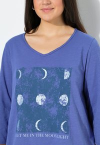 Langarm, diep V-hals top in effen blauwe katoen. De voorkant heeft een donkerblauw grafisch ontwerp met maanfasen en de tekst "Meet Me in the Moonlight."