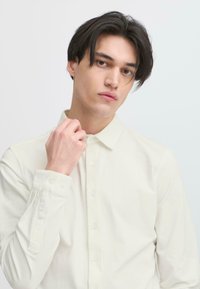 Light cream button-up shirt with a collar and long sleeves, featuring a smooth texture and subtle button accents. Model is adjusting the collar.