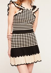 Stickad klänning med volangärmar i svart och krämfärgad, rutigt mönster, tätt liv och utsvängd kjol med scallopad fåll. Strukturerad tyg.