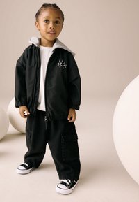 Veste noire zippée avec capuche grise, t-shirt blanc, pantalon cargo noir ample, et baskets noires avec accents blancs. Graphique simple de soleil sur la veste.