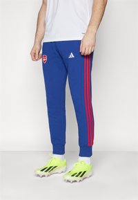 Modré tepláky Adidas s červenými pruhy, se znakem Arsenalu. Materiál vypadá měkce a silueta je u kotníků zúžená.