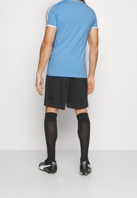 Muž v modré košili s krátkým rukávem, černých šortkách, dlouhých černých ponožkách a černých kopačkách, stojí čelem k bílé zdi.