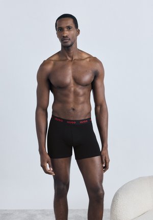 Μαύρα ανδρικά μποξεράκια με κόκκινο λογότυπο "HUGO" στην ελαστική μέση, κατασκευασμένα από λείο, ελαστικό ύφασμα. Η εφαρμογή είναι στενή στο σώμα, με μινιμαλιστικό σχεδιασμό.