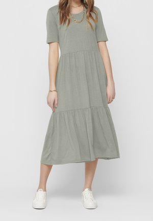 Femme portant une robe midi verte sauge à volants avec manches courtes, des baskets blanches et un collier et un bracelet en chaîne dorée.