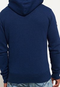 Hombre con una sudadera azul marino sencilla y jeans azul claro, mostrado de espaldas contra un fondo blanco.