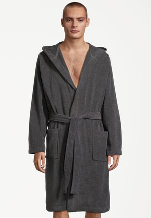 Młody mężczyzna w ciemnoszarym szlafroku z kapturem, paskiem i dwoma dużymi przednimi kieszeniami, stojący na białym tle.