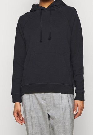 Personne portant un sweat à capuche noir avec poche avant et cordons, associé à un pantalon gris ample, sur un fond uni.