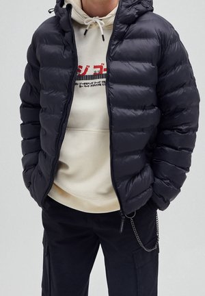 Homme portant une veste matelassée noire par-dessus un sweat à capuche crème avec un motif rouge, un pantalon noir avec des poches latérales, et un accessoire chaîne sur le côté droit.