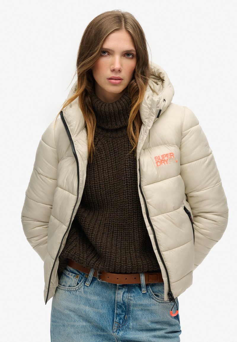 Beige Steppjacke mit gesteppter Optik, Reißverschluss und Kapuze. Das Logo auf der Brust in hellem Korallenrot ist zu sehen, getragen über einem strukturierten braunen Pullover.