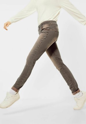 Henkilö, jolla on käärityt ruskeat slim fit -farkut, valkoiset sukat, valkoiset lenkkarit ja pitkähihaisen vaaleanbeigen paita, kävelee kesken askelen yksiväristä taustaa vasten.