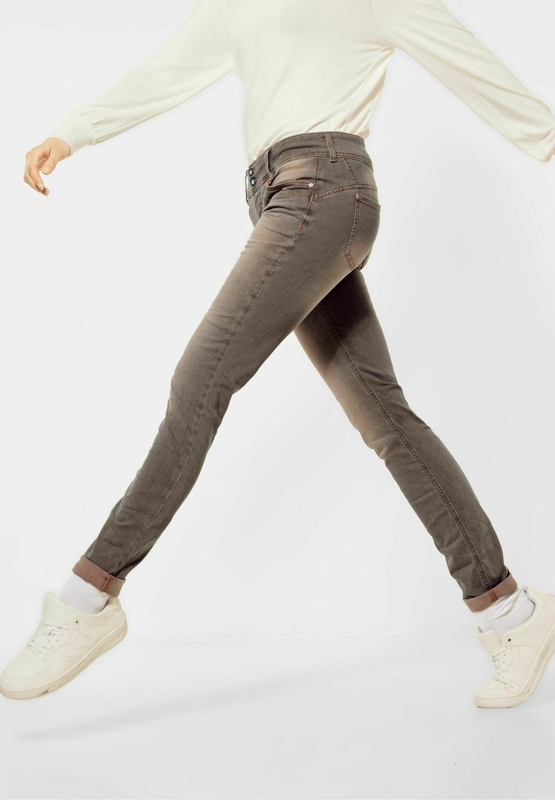 Personne portant un jean slim marron retroussé, des chaussettes blanches, des baskets blanches et un haut beige clair à manches longues, en plein milieu d'une foulée sur un fond uni.